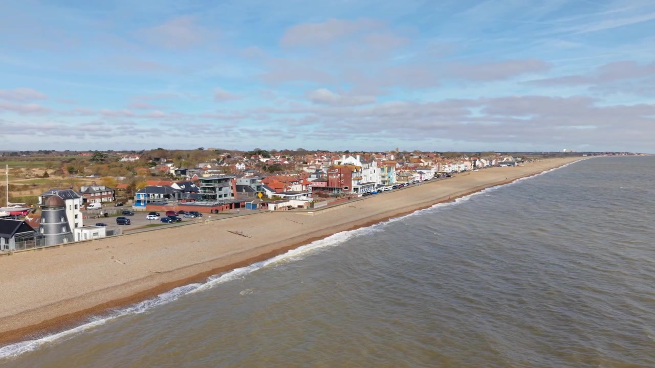 Verhoogd dronebeeld langs de kustlijn van Aldeburgh met pastelkleurige huizen en brekende golven.