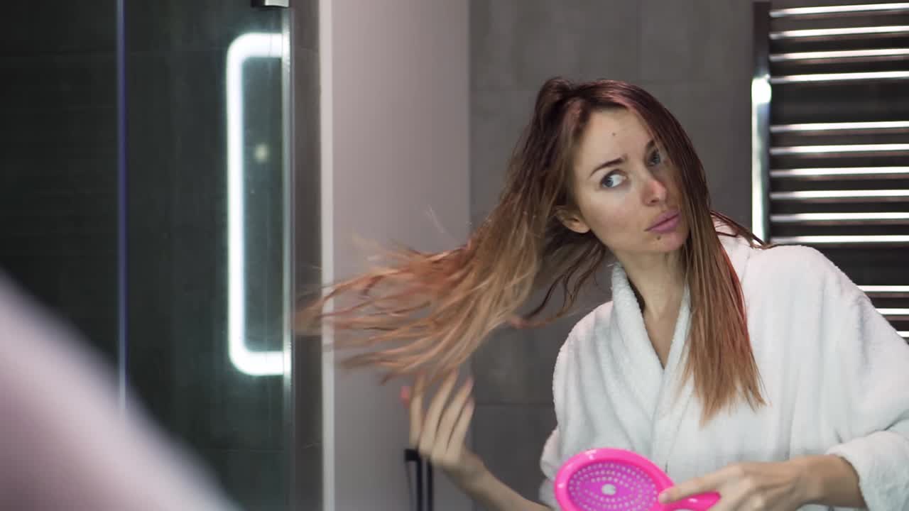 Mujer bonita peinándose el cabello largo mirándose al espejo; chica cepillándose el pelo en el baño.