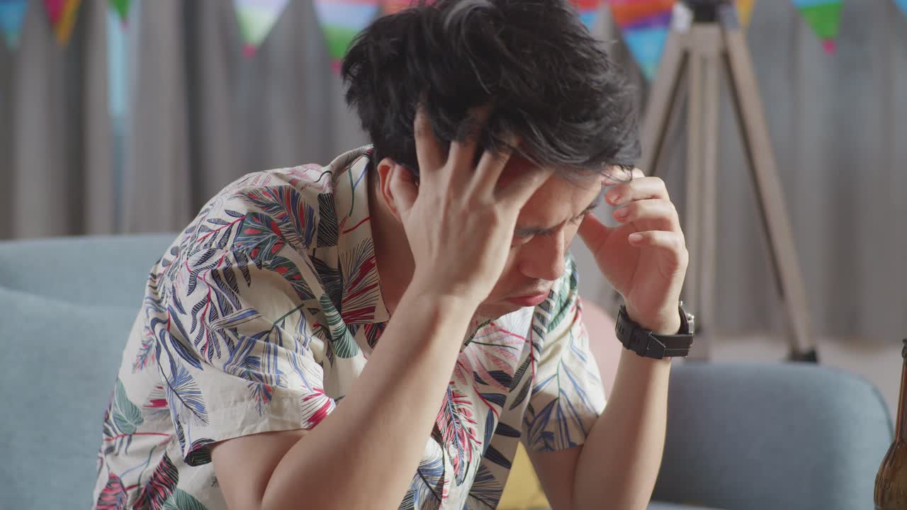 소파에 앉아있는 술에 취한 아시아 남자가 집에서 파티 방에서 두통으로 인해 머리를 손으로 잡고 있습니다.