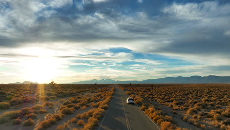 Eine-Einsame-Straße-Durch-Die-Mojave-wüstenlandschaft,-Während-Ein-Einsames-Auto-Bei-Sonnenuntergang-Die-Straße-Hinunterfährt---Ziehen-Sie-Die-Luftaufnahme-Zurück