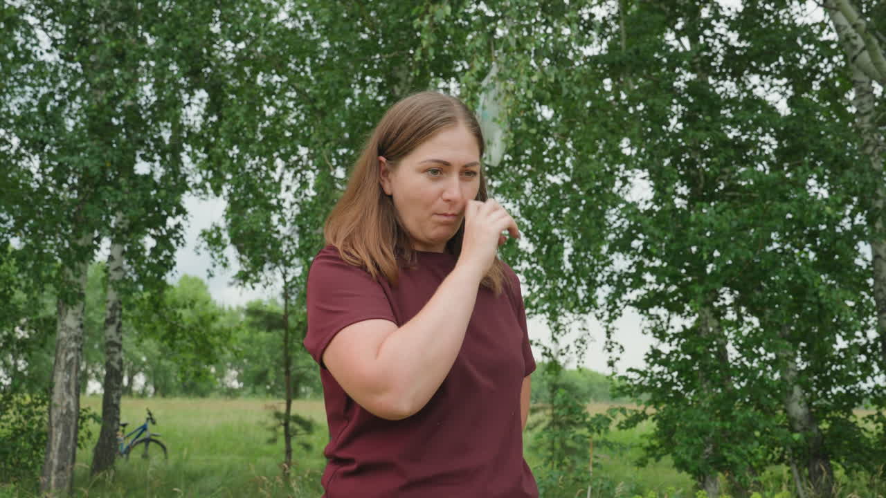 Mujer blanca probando una baya silvestre madura al aire libre; recolectora prueba una baya cerca de abedules, con camisa informal color burdeos, primer plano de la mano a la boca, sereno prado de verano, expresión sincera de curiosidad, ambiente rural tranquilo