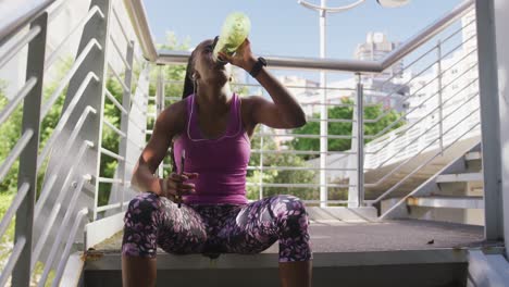 african american woman drinking water and using smartphone sitting on the stairs of the city bridge