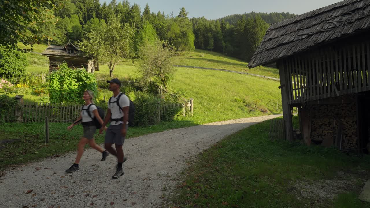 A young couple hikes along a quiet rural path surrounded by green fields and wooden cabins