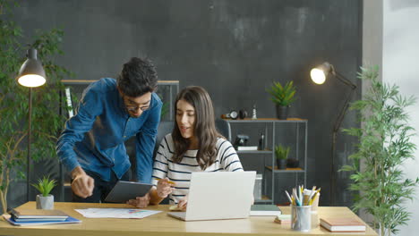 Arabischer-Mann-Und-Brünette-Frau-Sitzen-Zusammen-Am-Schreibtisch,-Arbeiten-Und-Reden-Vor-Dem-Laptop-Im-Büro