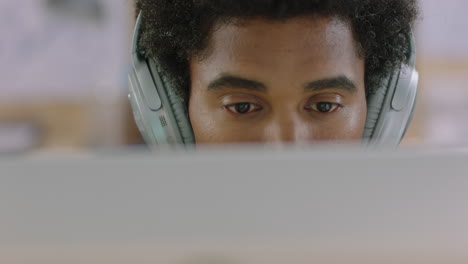 Joven-Hombre-De-Negocios-De-Raza-Mixta-Usando-Una-Computadora-Trabajando-En-Un-Proyecto-De-Negocios-Navegando-En-Línea-Escuchando-Música-Intercambiando-Ideas-Usando-Audífonos-En-Un-Lugar-De-Trabajo-De-Oficina-Relajado