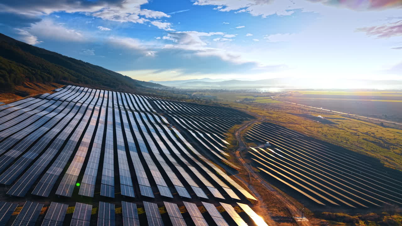 Solcellepark i den gyldne time ved solnedgang. Luftperspektiv af en solenergipark med skarp sollysrefleksion og linseflare over en bjergdal.