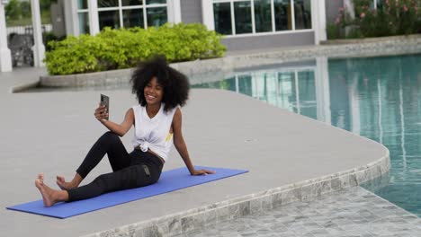 a female dressed for exercise sits on a yoga mat talking smart phone video call.