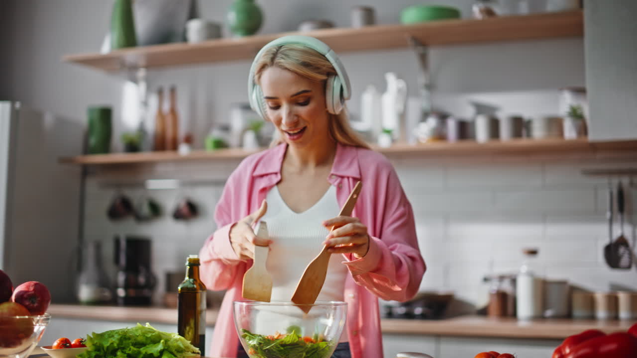 Headphones girl cooking meal dancing joyfully at kitchen. Woman pouring oil