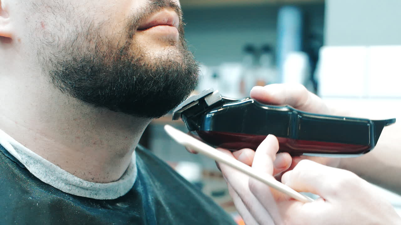 Hairdresser cutting beard to client with clipper. Close up of young bearded man