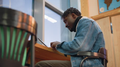 young male visitor sits at wooden desk near window scrolling on tablet wearing denim jacket and turtleneck inside modern bright coworking or library space focused posture natural daylight illumination