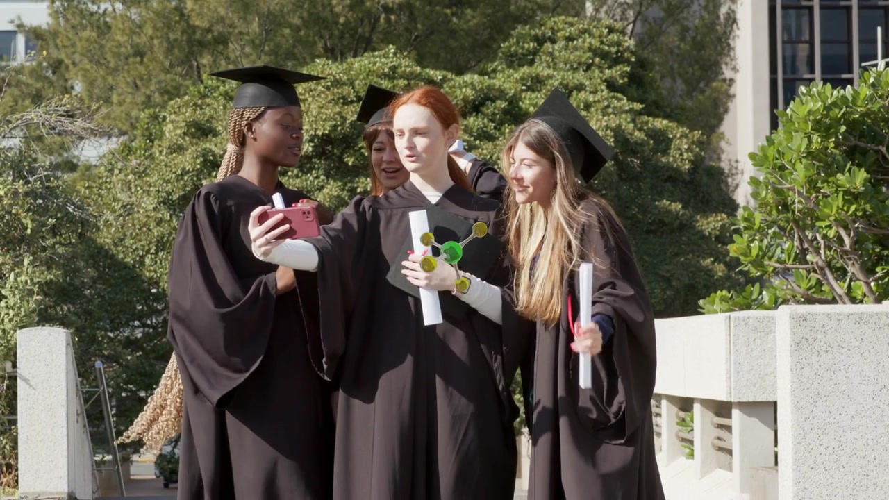 Quattro laureati che salgono i gradini, la donna in testa alza il telefono, molecole che fluttuano sullo schermo del telefono per la laurea