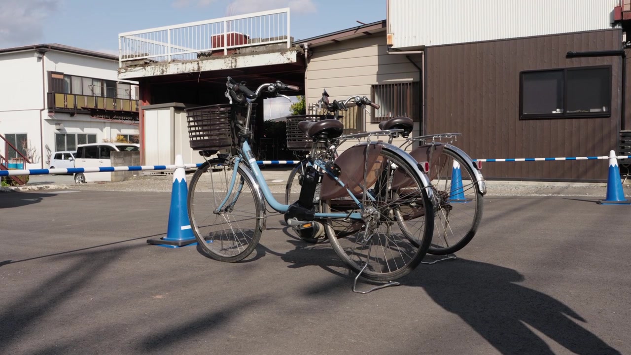 日本の郊外の通りで、青いパイロンの横のアスファルトに駐輪されたカゴ付きの自転車2台。