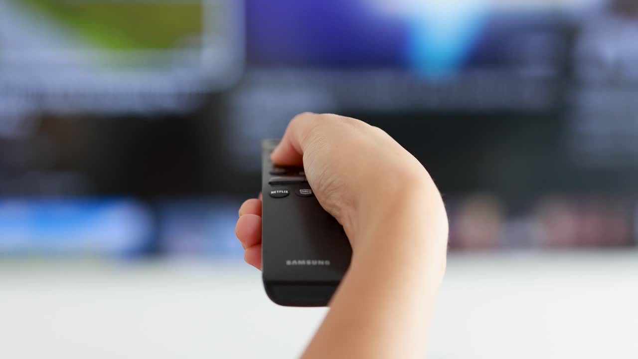 A person’s hand holds a remote control, pointing at a television in a bright indoor setting, pressing buttons to switch channels. Shallow depth of field, steady camera