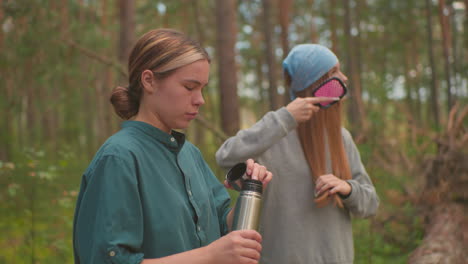 Side view of sisters pause in tranquil forest, one opens thermos by pressing inner cover, while other gently brushes her hair, backpacks rest on fallen tree as they enjoy serene moment together