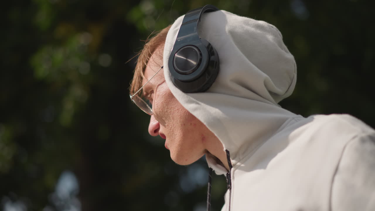 joven con auriculares en el exterior, retrato con sudadera en un parque urbano, estudiante pensativo disfrutando de una banda sonora lofi bajo una luz suave, primer plano del perfil y ángulos espontáneos que capturan una expresión contemplativa