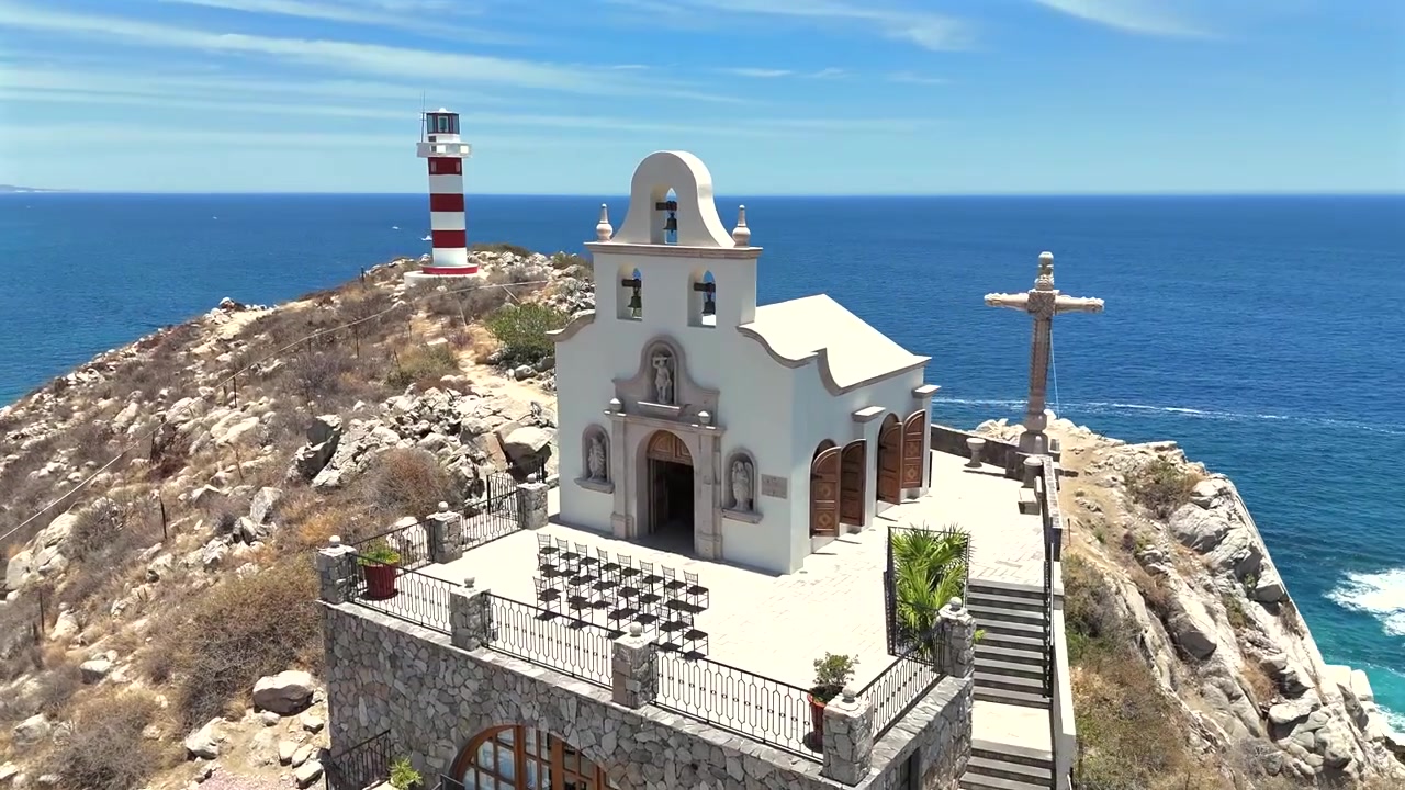 Un drone si allontana in orbita dalla storica chiesa sulla scogliera e dal Faro El Encanto a Cabo San Lucas, allontanandosi per rivelare l'oceano aperto e la spiaggia che si estende sotto.