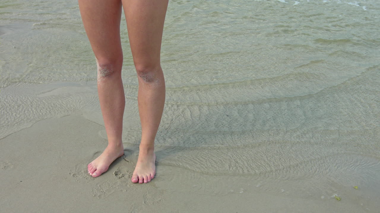 tiro estático piernas deportivas largas y femeninas de pie en la orilla del mar de la playa, cámara lenta