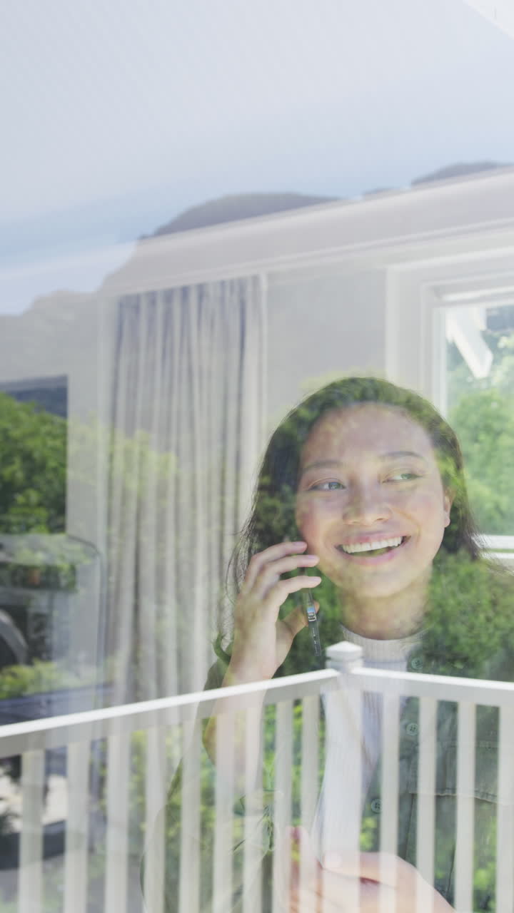 Vertical video of happy asian woman looking through window and using smartphone, in slow motion