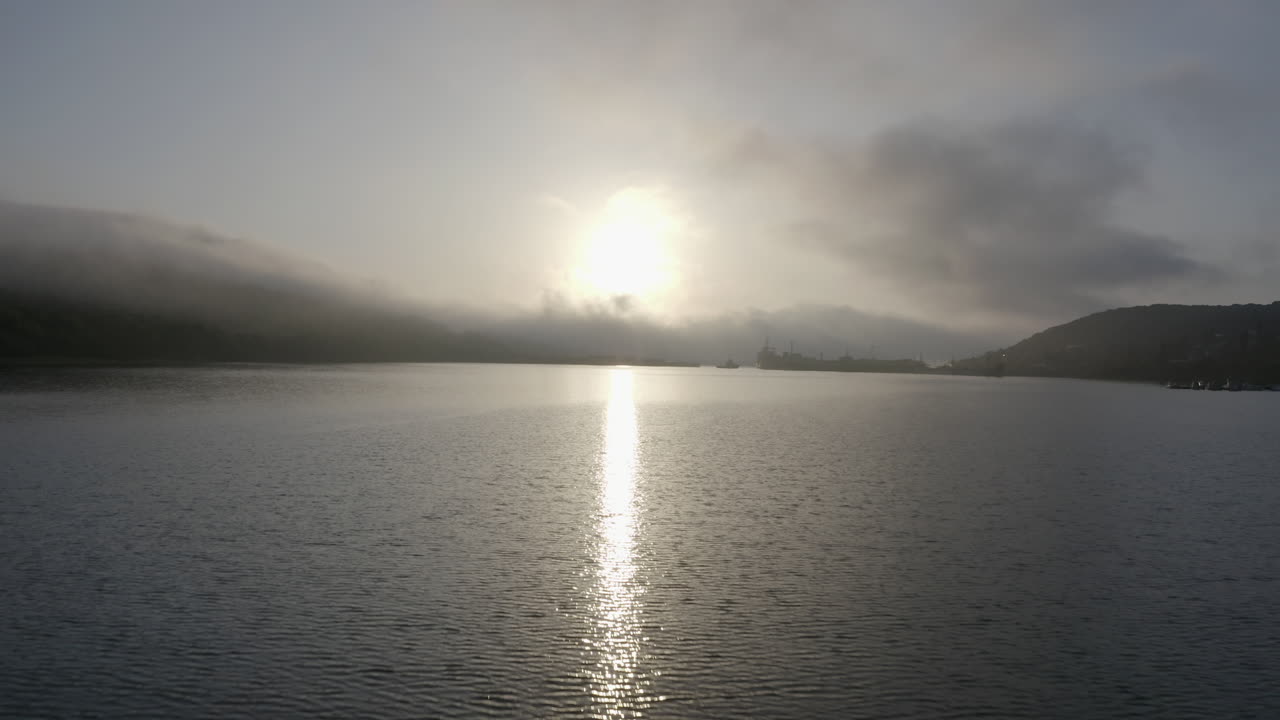 만의 물 표면 위로 가까이 날아가며, 해가 물 표면에 반사되고, 언덕 위에서 오는 밀집한 안개 구름, 해가 지는