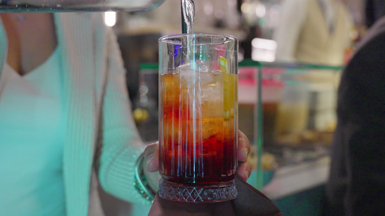 Bartender pours layered cocktail into a tall glass at a vibrant indoor bar scene