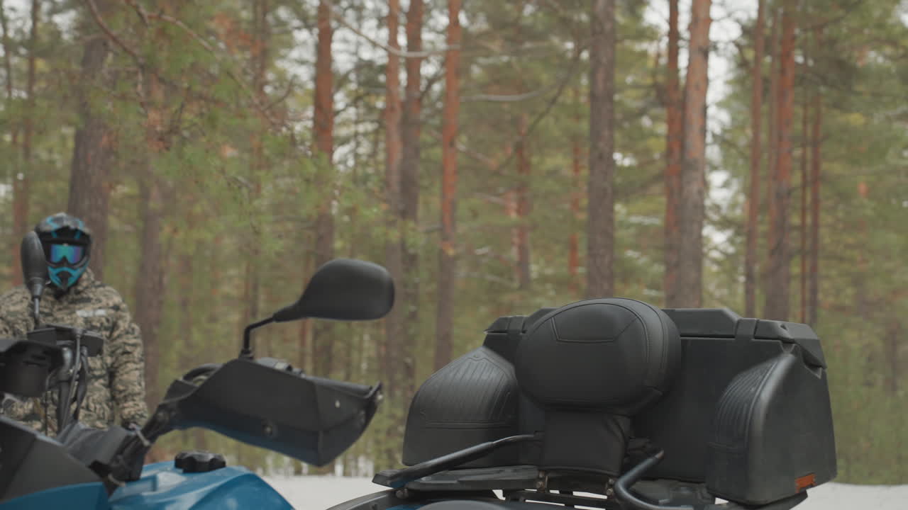 Preparazione dell'ATV in pineta innevata: il pilota controlla casco ed equipaggiamento vicino a un quad mimetico con occhiali rossi, scatto di profilo ravvicinato, momento di silenziosa concentrazione, fiato freddo nell'aria, attesa prima dell'off-road estremo.