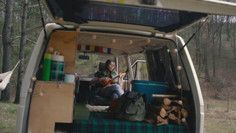 a young girl with red hair plays the ukulele inside a caravan in the countryside