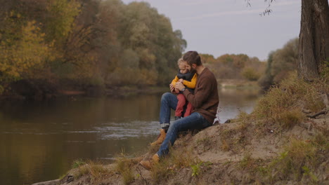 trip-in-forest-park-at-autumn-day-young-father-and-little-child-are-sitting-on-coast-of-small-river-resting-at-nature-at-good-fall-weather