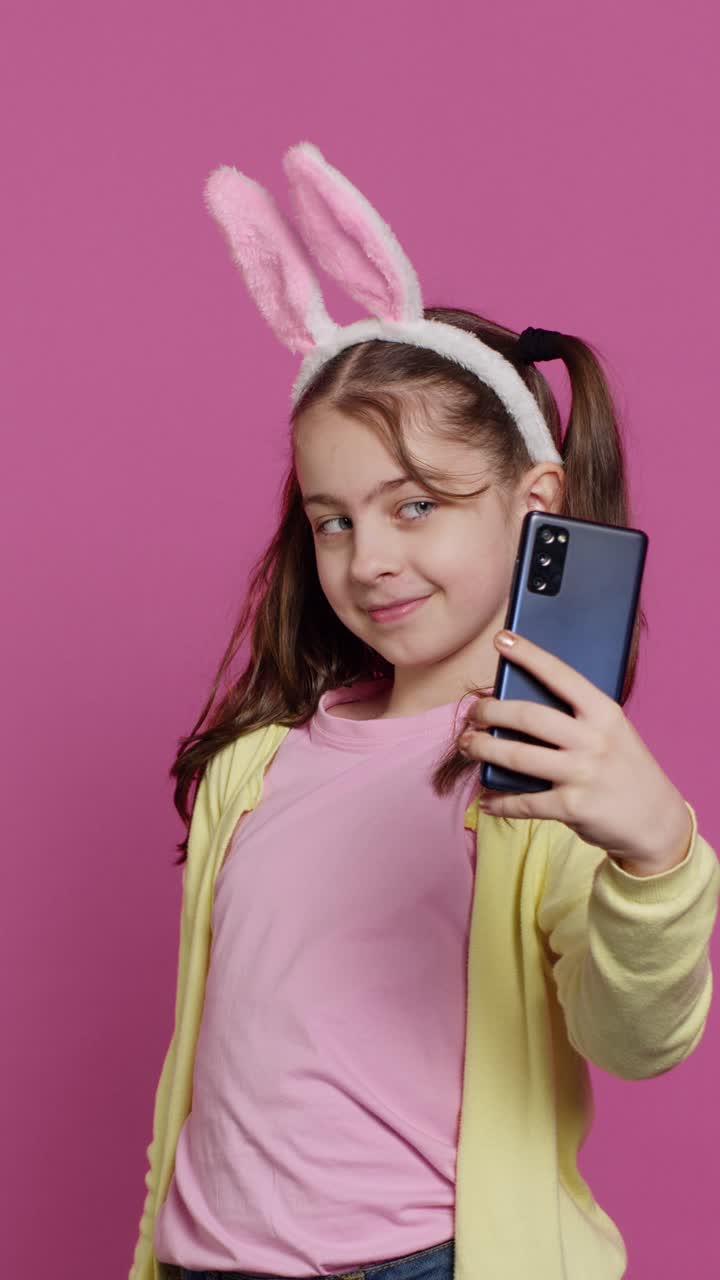 Bambina allegra con orecchie da coniglietto che si fa dei selfie su sfondo rosa