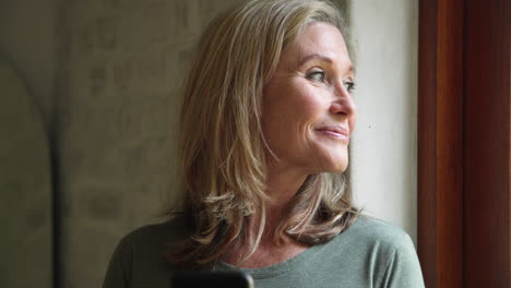smiling senior woman looking out window, holding smartphone at home
