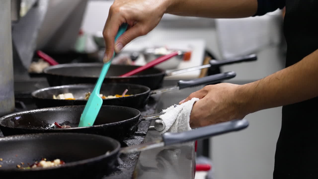 Restaurant cook stirs pan of food on stovetop alongside multiple skillets of cooking food, slider slow motion 4K