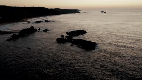 aerial tilt up: silhouette of rocky coastline and mountains with tranquil pacific ocean during sunset in backdrop