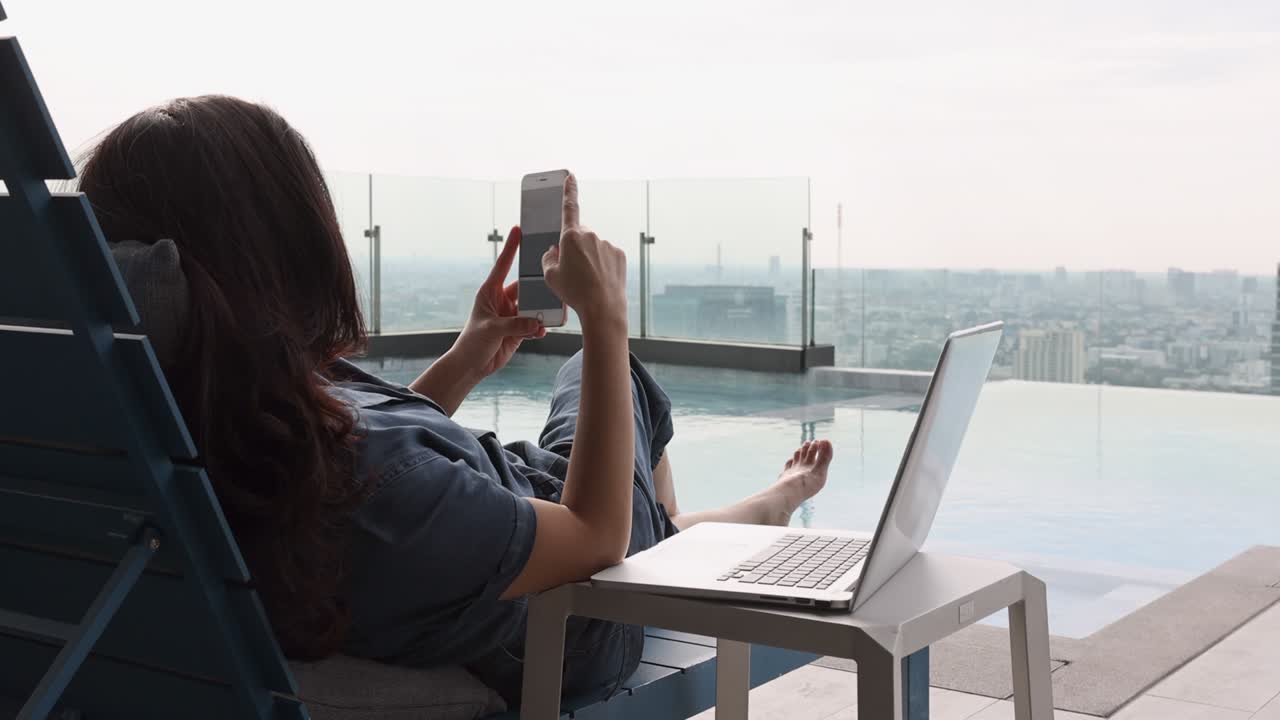 día de verano estilo de vida mujer asiática relajarse y relajarse cerca de la piscina de lujo. mujer asiática usando aplicación en un teléfono inteligente y trabajando en una computadora portátil y fondo borroso rascacielos, hermoso cielo y nubes.