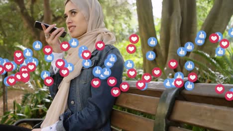 heart and like icons against woman in hijab talking on smartphone sitting on a bench in the park