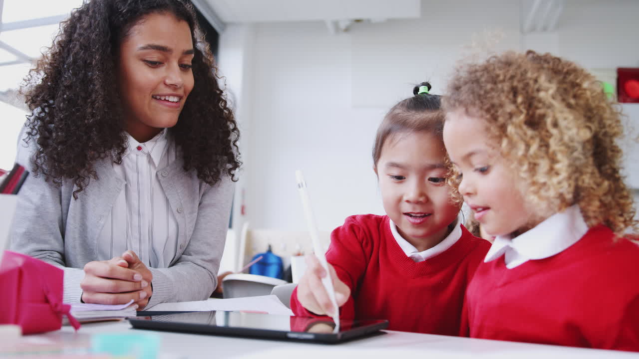 Maestra della scuola dell'infanzia che aiuta due bambine a usare un tablet e una penna stilo in classe, primo piano.
