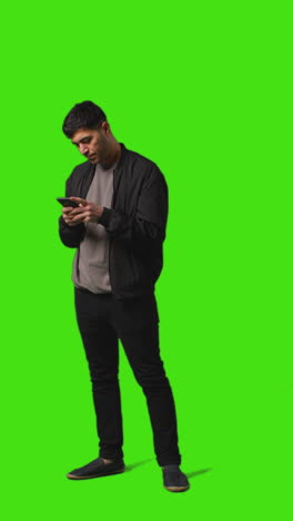 video vertical de larga duración de un joven que envía mensajes de texto en un teléfono móvil de pie contra una pantalla verde 1