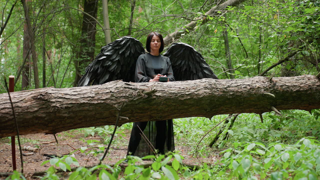 Female black witch with large dark wings typing on small device in forest, standing behind fallen tree surrounded by lush green foliage, appearing thoughtful and mysterious in magical woodland