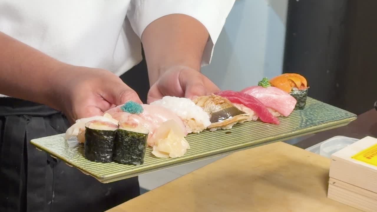 明るい室内照明の下、緑色の皿に盛り付けられたさまざまな握り寿司を、シェフが丁寧に盛り付けている。