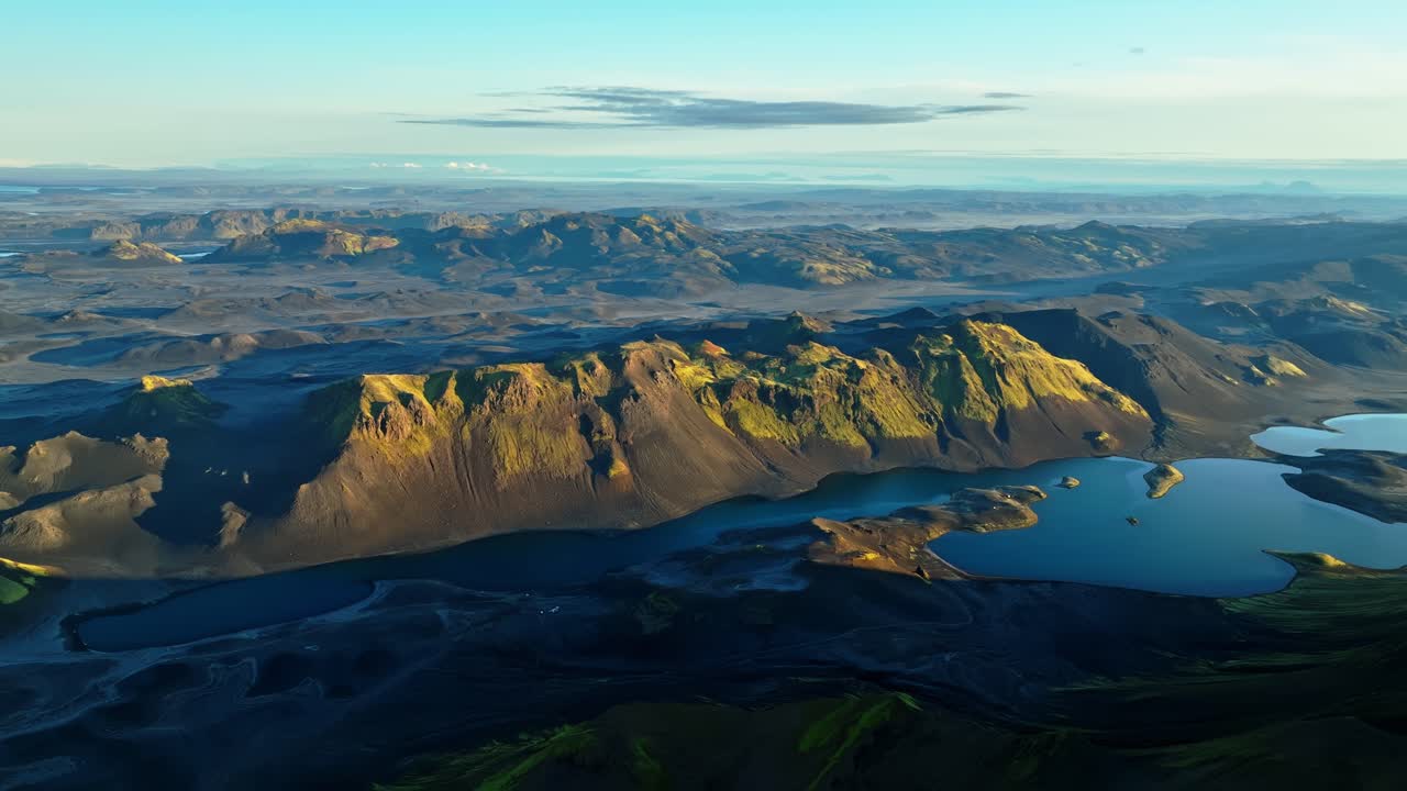 Dronepanorama av Islands høyland i den gylne timen, solbelyst vulkansk fjellrygg og svingende innsjø med øyer, mørke lavasletter og fjerne fjell som forsvinner i horisonten