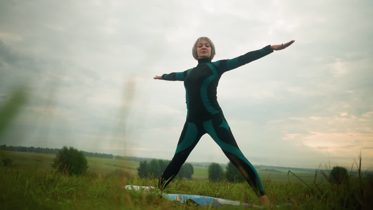 녹색과 검은색 의상을 입은 여성이 요가 매트 위에 서서 팔과 다리를 고 조용하고 잔디에 구름이 가득한 하늘을 배경으로 요가 삼각형 자세를 연습하고 있습니다.