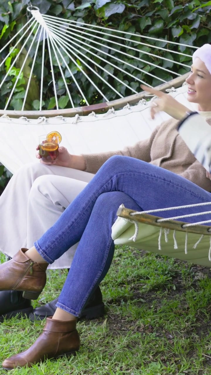 Video verticale: Con panoramica, donna con cappello di Babbo Natale che tiene una bevanda sull'amaca, indicando il giardino.