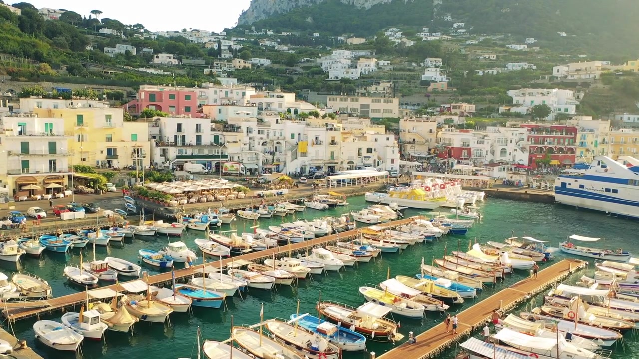 Ripresa aerea con drone del porto di Marina Grande in Grecia, pieno di barche bianche e traghetti vicino a edifici colorati e colline terrazzate che creano una vivace scena di città costiera.