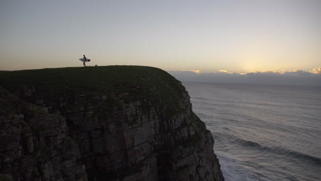 Man-with-surfboard-on-cliff-at-sunrise-by-ocean