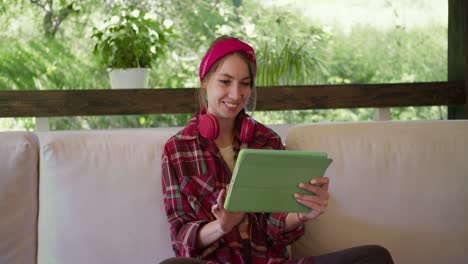una chica rubia en una camisa a cuadros roja con auriculares rojos y un vendaje rojo se sienta y trabaja en una tableta verde en un sofá en un gazebo en la naturaleza
