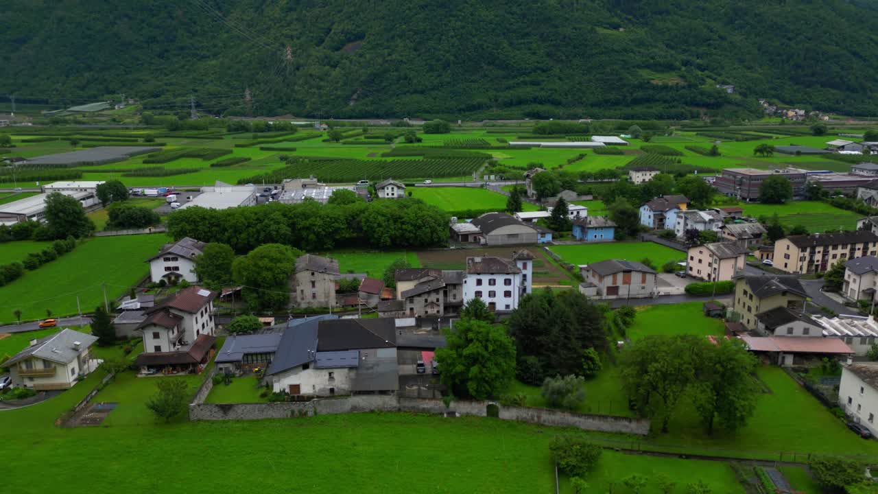 Drone shot of an Italian village surrounded by bright green fields and foothills in the Alpine valley. Shot in Villa di Tirano, Sondrio, Italy (Italia)
