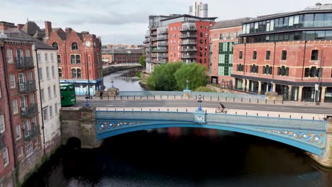 a green double-decker bus travels across a blue victorian bridge over the river aire, surrounded by historic red-brick buildings under overcast daylight