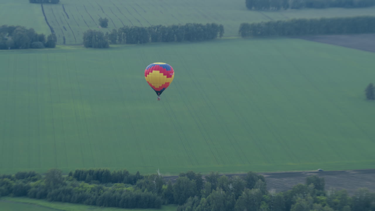 Distant view of colorful hot air balloon drifting over expansive green farmland, silhouette against fields and tree lines as gentle breeze carries vessel across serene tranquil landscape