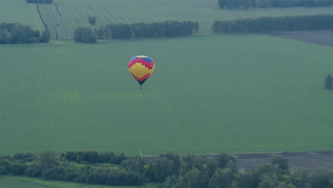 distant view of colorful hot air balloon drifting over expansive green farmland, silhouette against fields and tree lines as gentle breeze carries vessel across serene tranquil landscape