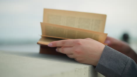 close up hand view of an individual in grey clothing holding an open book with their right hand on the pages as the wind softly flips through the book, the background is blurred