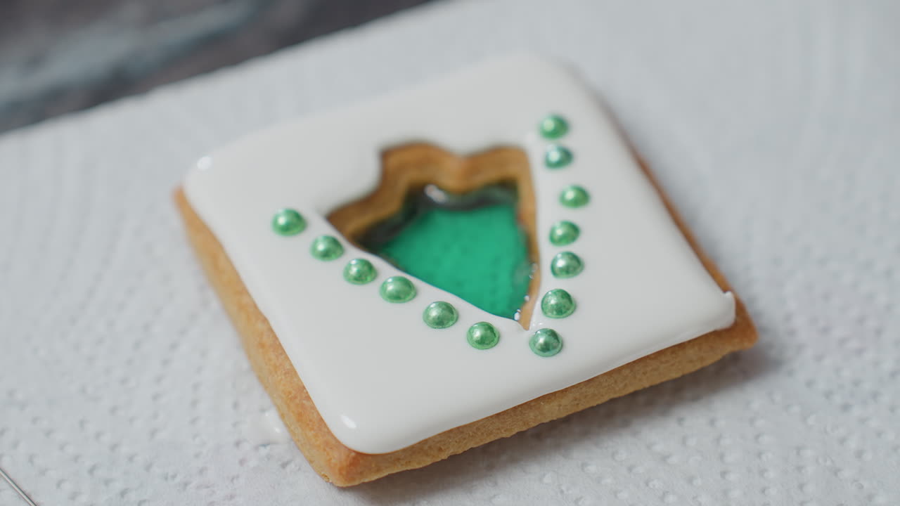 Close-up of beautifully decorated cookie with white icing and green pearl embellishments placed on tissue paper, the cookie has a cut-out center filled with a glossy green design