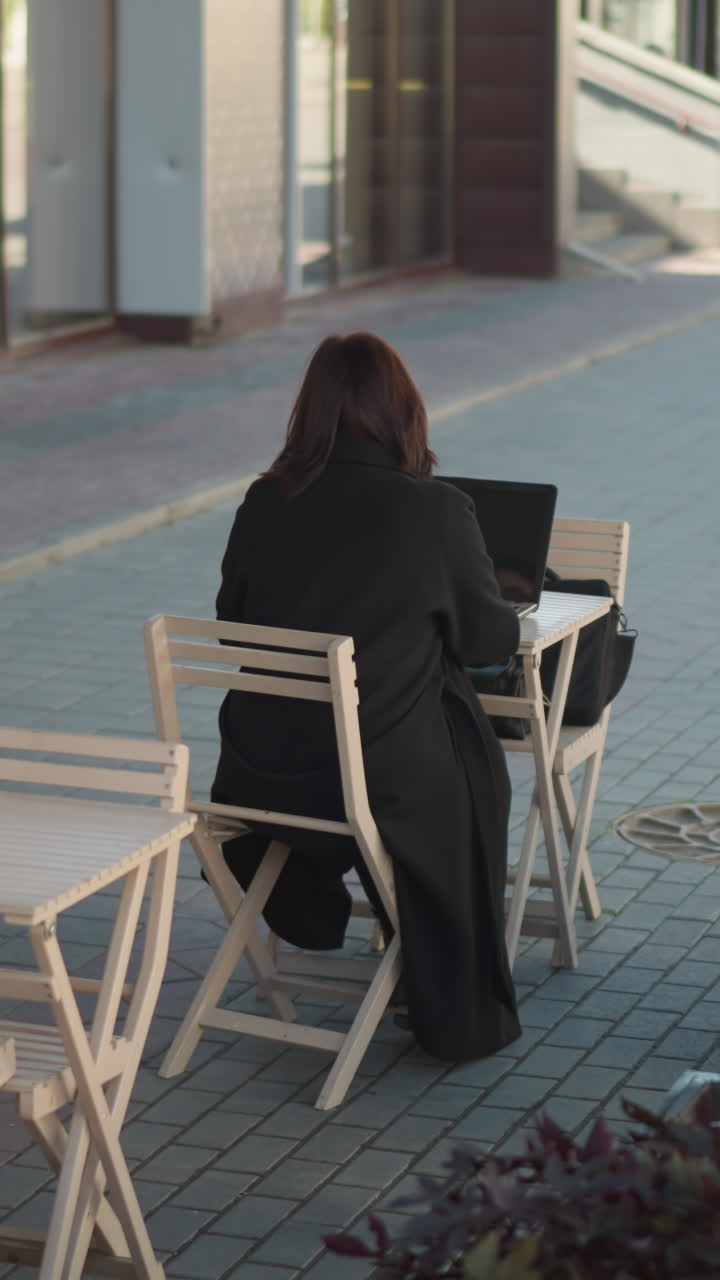 야외에서 노트북으로 타이핑하는 여자의 뒷면, 흑색 옷을 입은 사람이 멀리 걸어가고, 식물과 나무 의자가 주변에 있습니다.